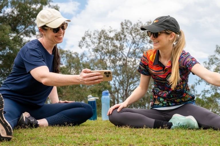 Two healthy women sitting stretching on the grass while looking at a phone together