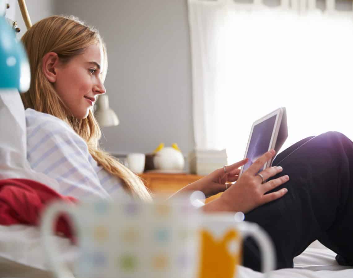 Young woman sitting on her bed, looking at a tablet device