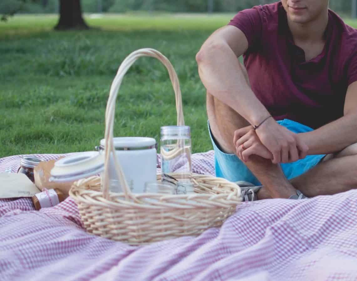 Man sitting on a picnic blanket beside a basket and food jars