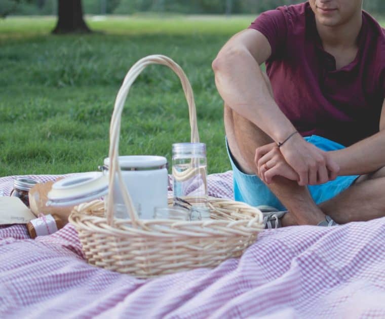 Man sitting on a picnic blanket beside a basket and food jars