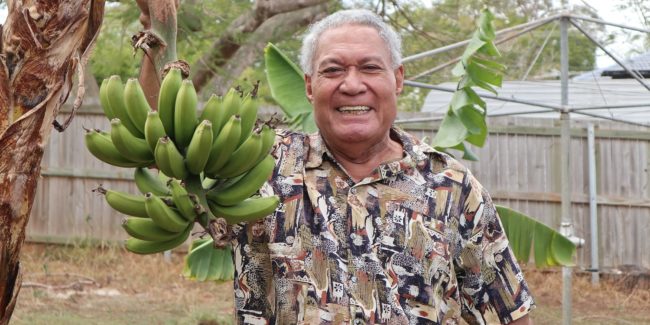 Man smiling while holding a banana bunch in his backyard