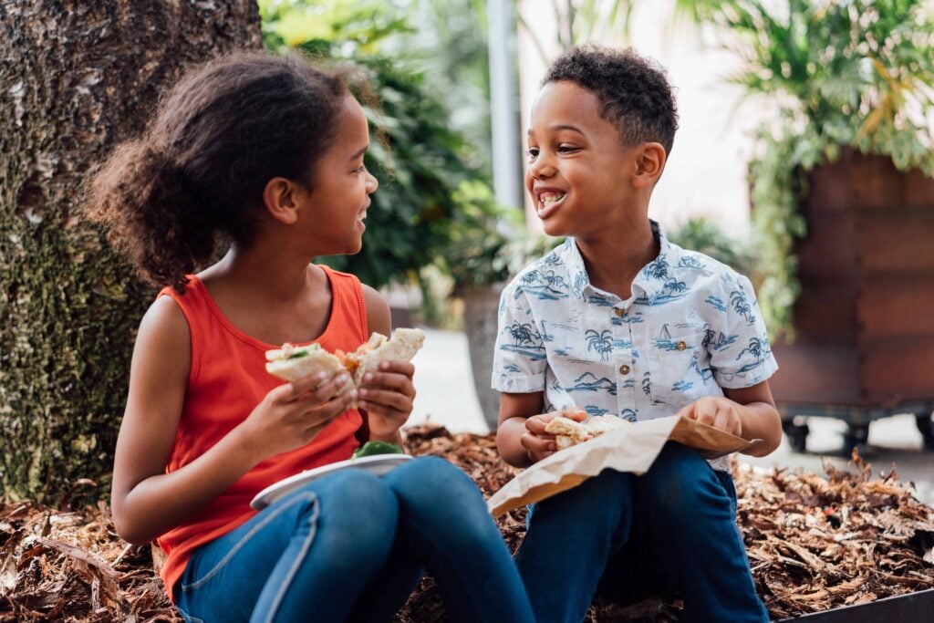 A young girl and boy are smiling at each other while eating healthy sandwiches outside