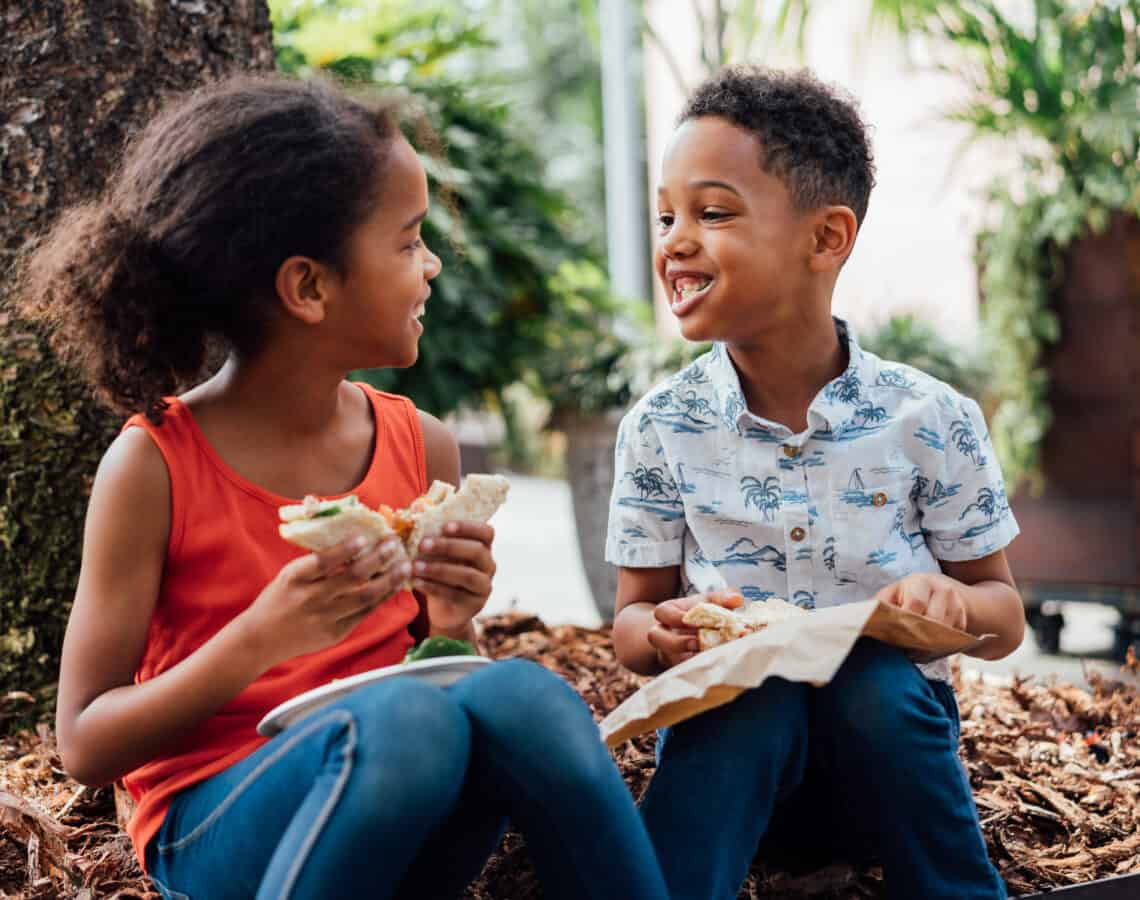 A young girl and boy are smiling at each other while eating healthy sandwiches outside