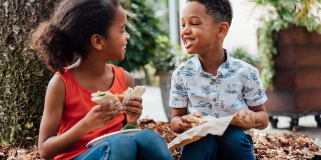 A young girl and boy are smiling at each other while eating healthy sandwiches outside