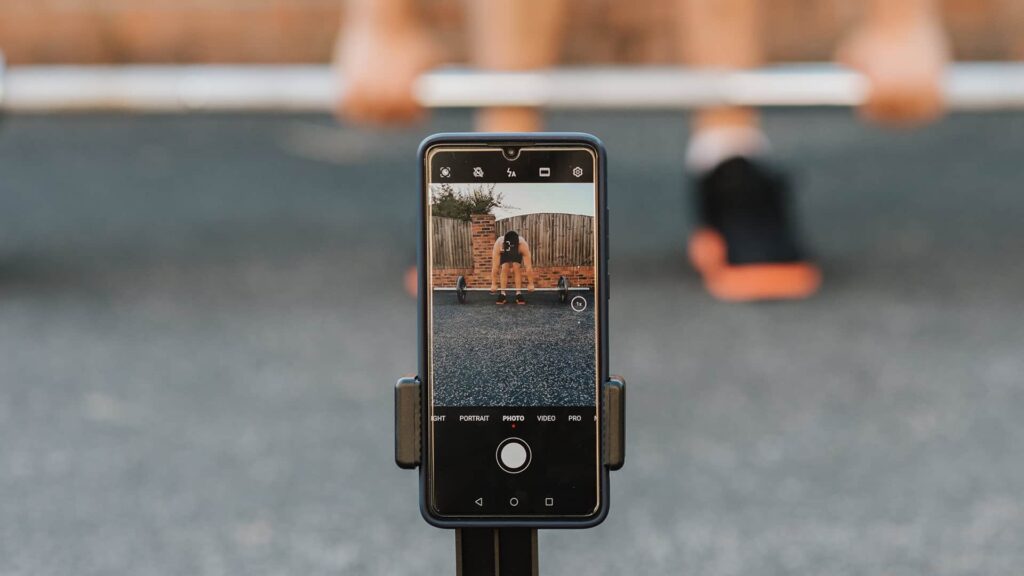 A man wearing sports shoes standing behind weights is recording his workout using a mobile phone