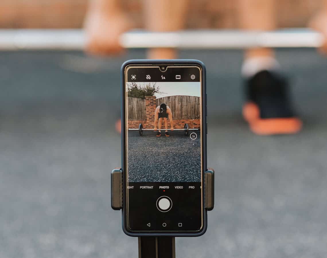 A man wearing sports shoes standing behind weights is recording his workout using a mobile phone