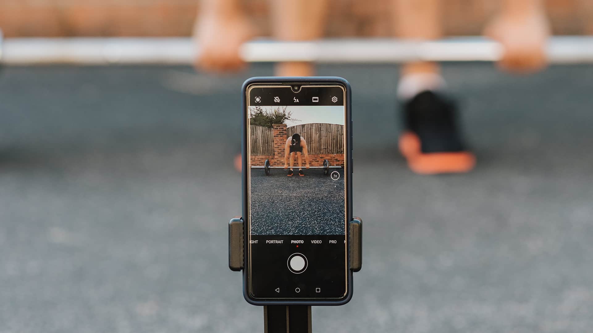 A man wearing sports shoes standing behind weights is recording his workout using a mobile phone