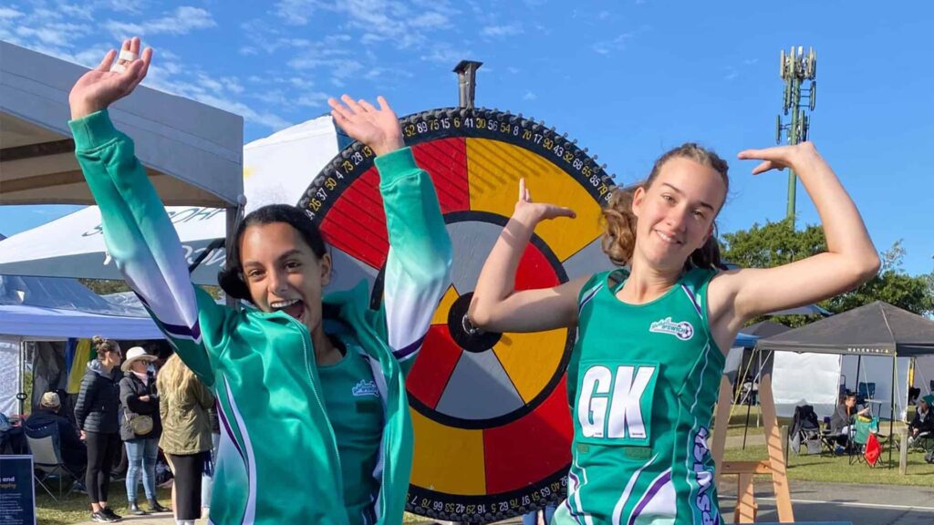 Netballers jump for joy beside the Health and Wellbeing Queensland prize wheel