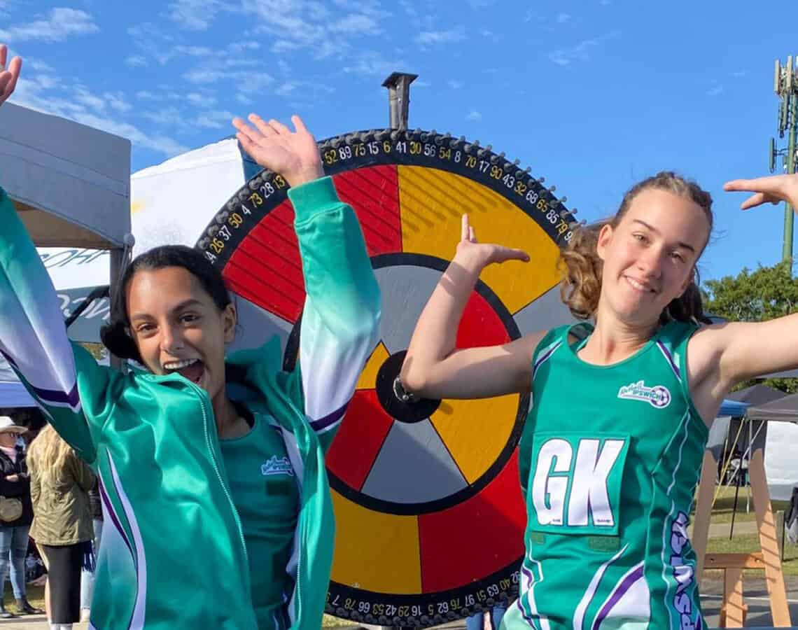 Netballers jump for joy beside the Health and Wellbeing Queensland prize wheel