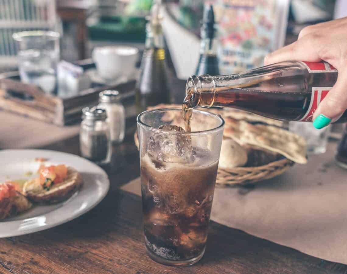 A glass of cola is being poured into a glass with ice that is sitting on a wooden table next to plates of bread and bruschetta