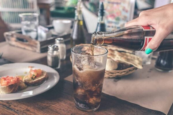 A glass of cola is being poured into a glass with ice that is sitting on a wooden table next to plates of bread and bruschetta