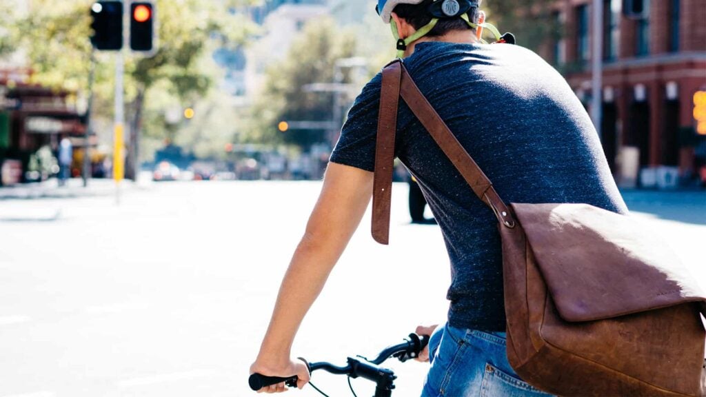 A person riding a bike who is wearing a helmet and stachel over their shoulder is stopped at a red traffic light on an urban road.