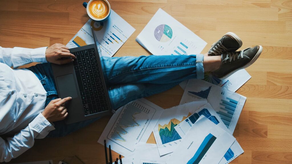 A person is sitting on the floor with a laptop on their lap. They are surrounded y papers and a cup of coffee.