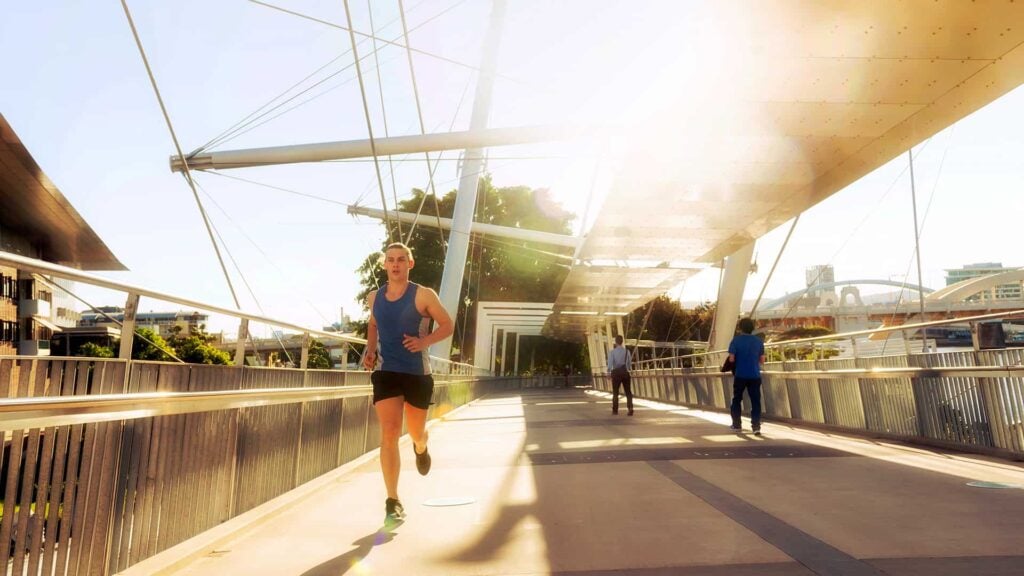A man wearing a blue top, black shorts and running shoes jogging across an urban bridge.