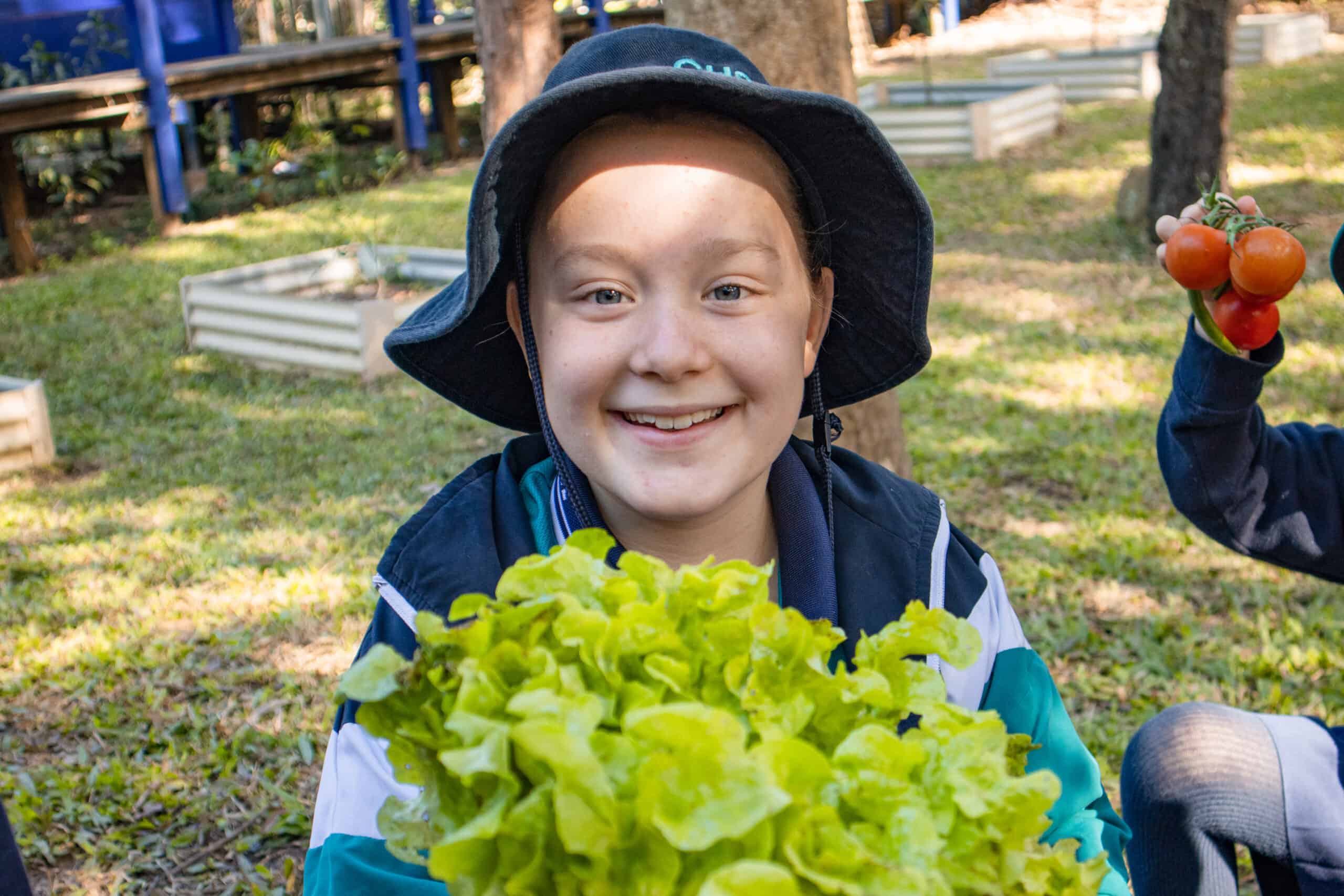 A young girl wearing a school uniform and hat holds a bunch of fresh lettuce