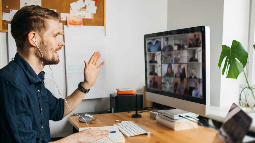 A man is sitting at a computer wearing headphones. He has a notebook and pen are in his hands. A video call with 16 people can be seen on the computer screen.
