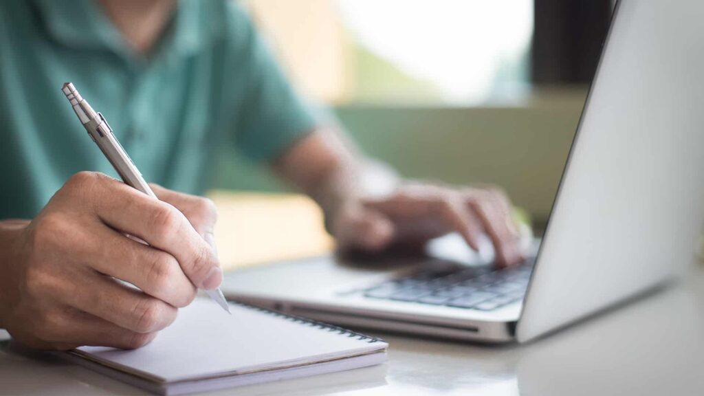 A person is sitting in front of an open laptop with a notebook and pen.