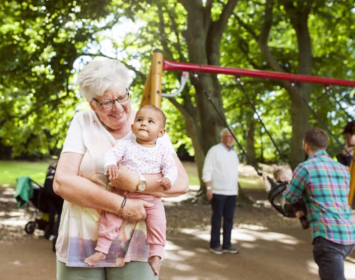 Grandmother carrying baby girl. Other families playing at a park behind them