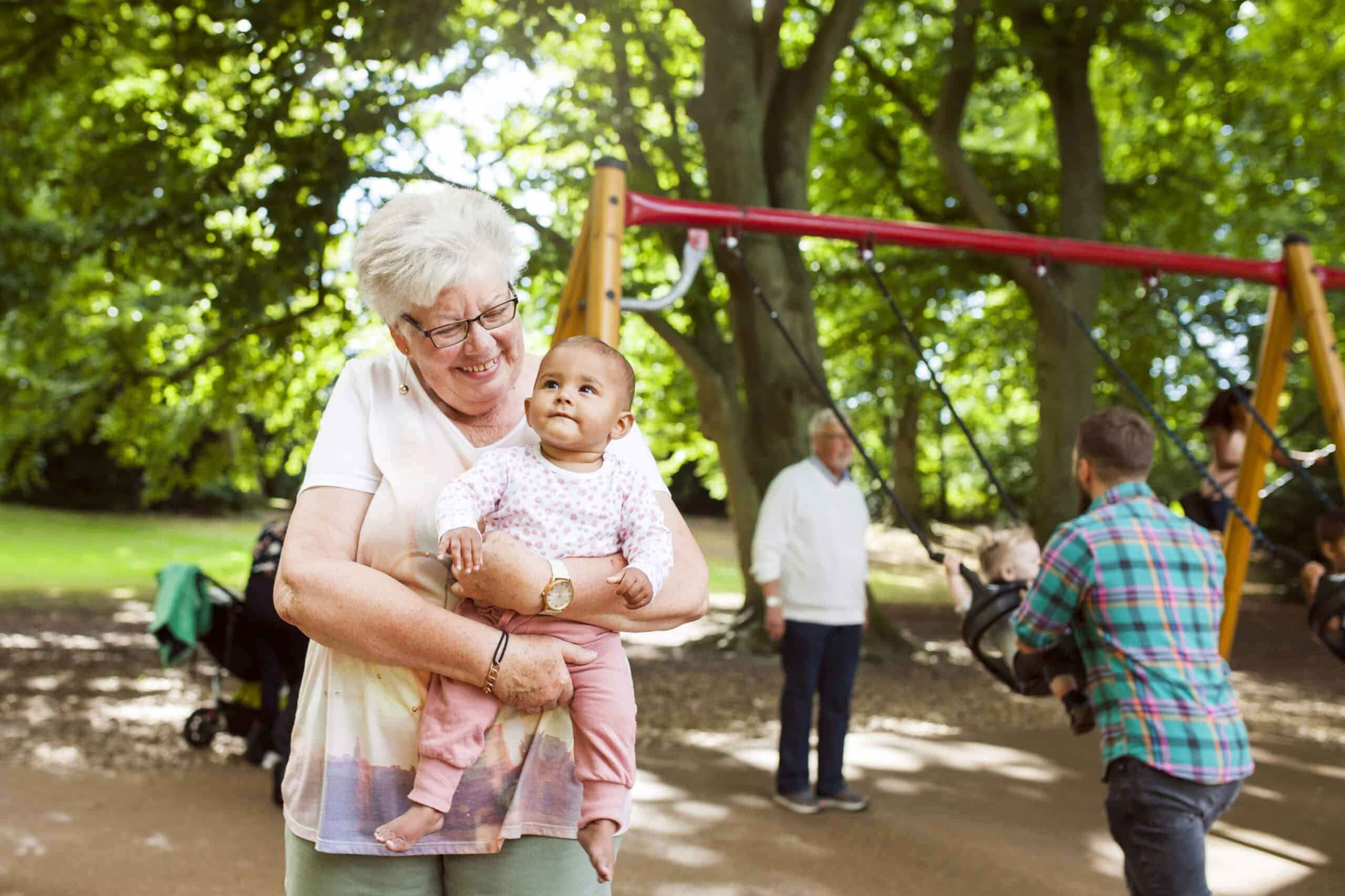 Grandmother carrying baby girl. Other families playing at a park behind them