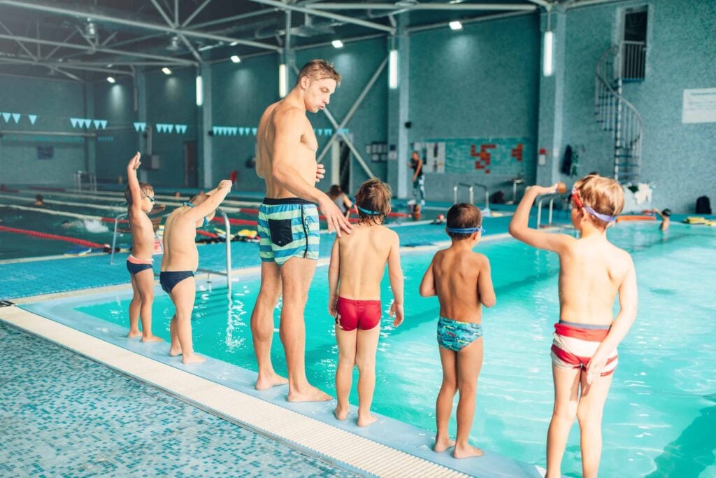 Instructor with children stands near water in pool and preparing for diving. Trainer shows kids exercise, view from back. Modern sports center on background. Sportive kids activity.