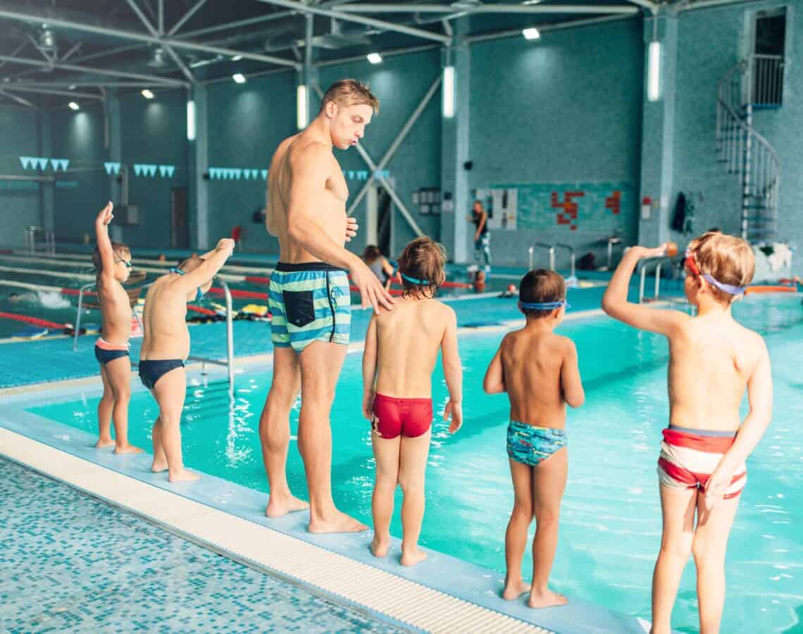 Instructor with children stands near water in pool and preparing for diving. Trainer shows kids exercise, view from back. Modern sports center on background. Sportive kids activity.