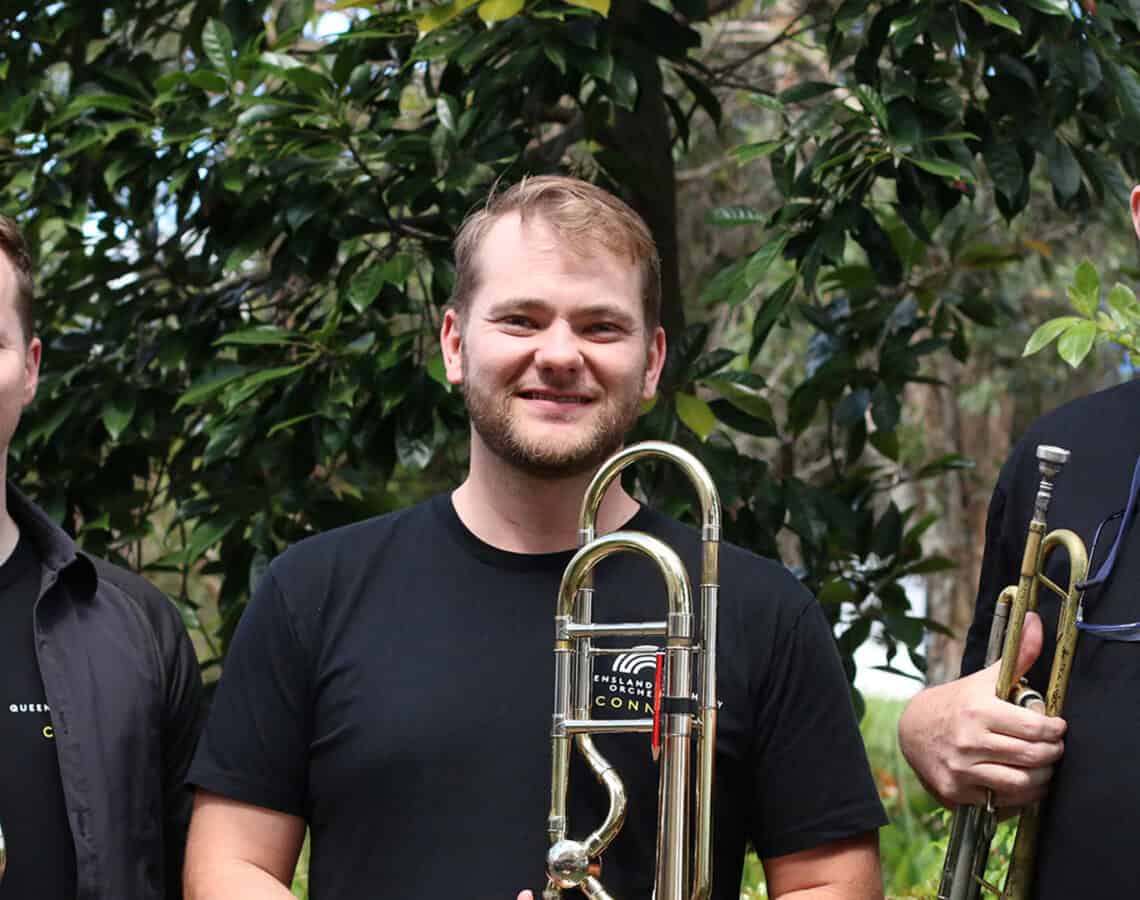 3 members of the Queensland Symphony Orchestra are standing outside holding brass instruments