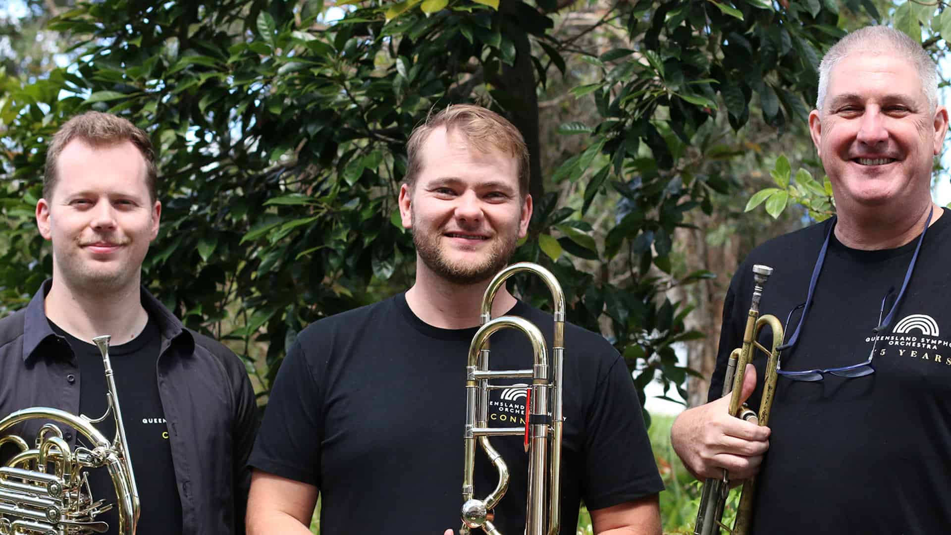 3 members of the Queensland Symphony Orchestra are standing outside holding brass instruments
