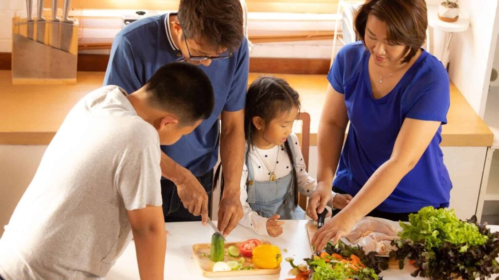 A family of 2 adults and 2 kids are preparing vegetables and a raw chicken in the kitchen.