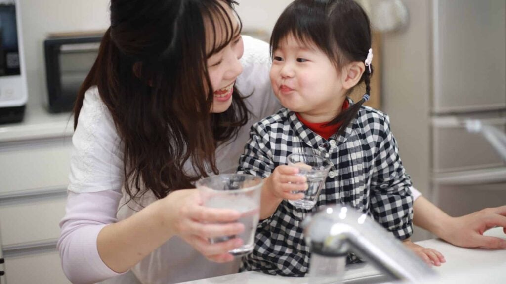 A mother and daughter drinking glasses of water behind a running kitchen tap.