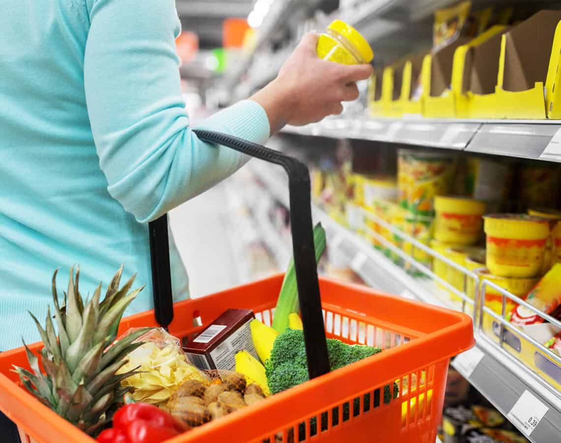 A woman wearing a blue top is holding a red shopping basket filled with groceries and looking at items in a supermarket aisle.