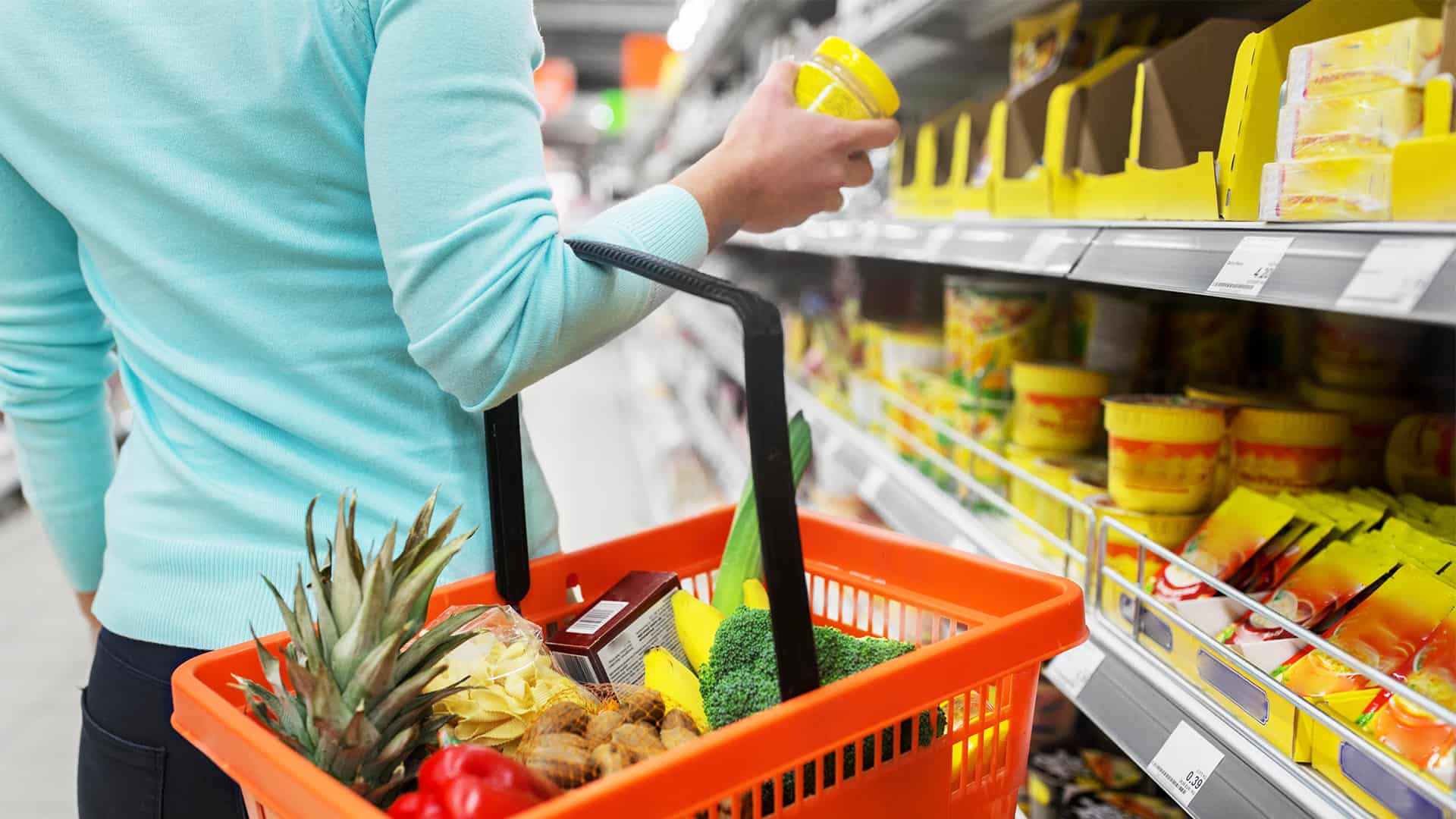 A woman wearing a blue top is holding a red shopping basket filled with groceries and looking at items in a supermarket aisle.