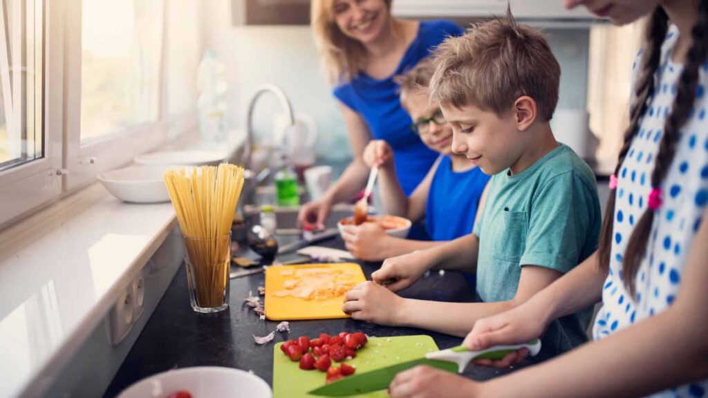 3 children cutting tomatoes and cheese on coloured cutting boards in a kitchen. A woman in a blue shirt is watching them and smiling. There is a glass of dry pasta and a bowl of pasta sauce on the bench.