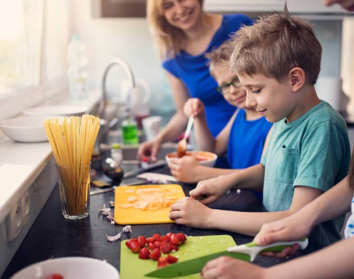 3 children cutting tomatoes and cheese on coloured cutting boards in a kitchen. A woman in a blue shirt is watching them and smiling. There is a glass of dry pasta and a bowl of pasta sauce on the bench.