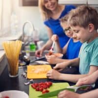 3 children cutting tomatoes and cheese on coloured cutting boards in a kitchen. A woman in a blue shirt is watching them and smiling. There is a glass of dry pasta and a bowl of pasta sauce on the bench.