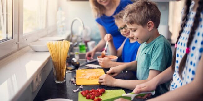 3 children cutting tomatoes and cheese on coloured cutting boards in a kitchen. A woman in a blue shirt is watching them and smiling. There is a glass of dry pasta and a bowl of pasta sauce on the bench.