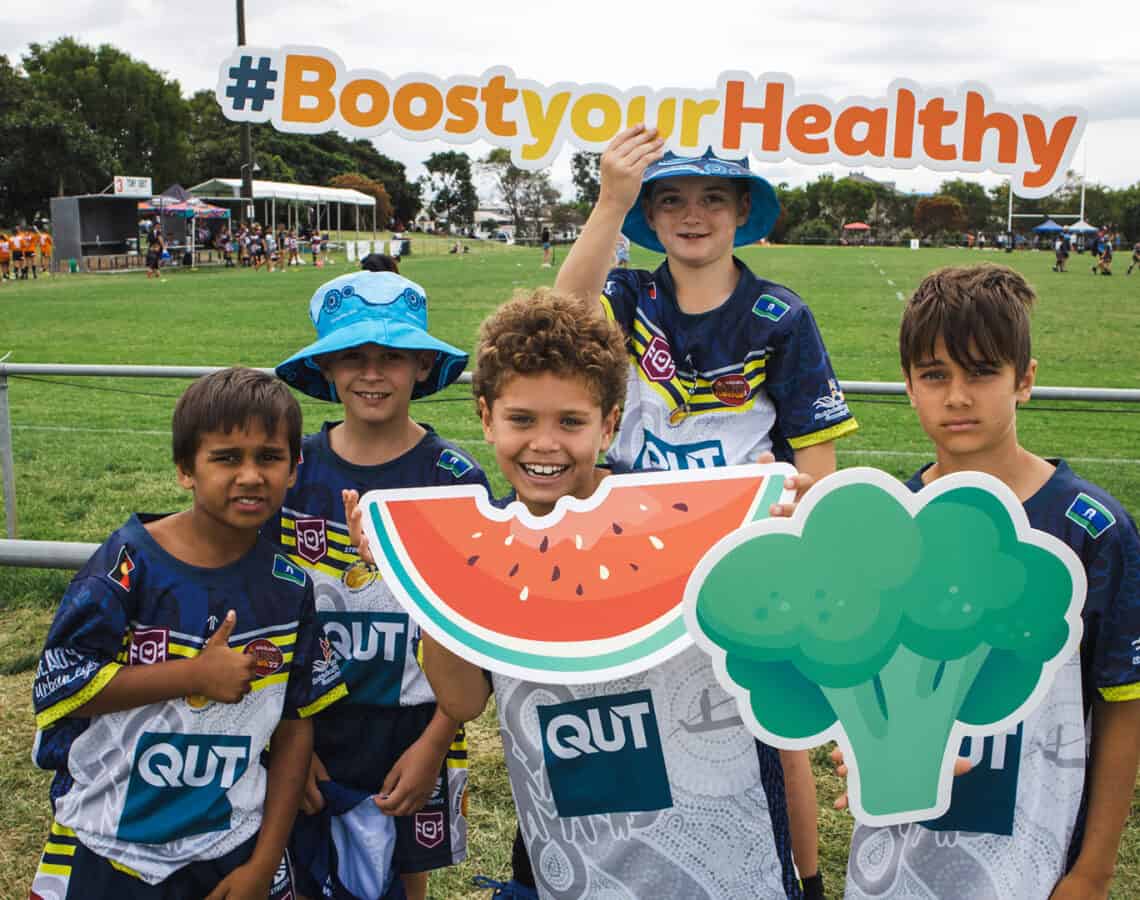 5 children on a sporting field are holding colourful signs in the shape of watermelon, broccoli and words that say #boostyourhealthy