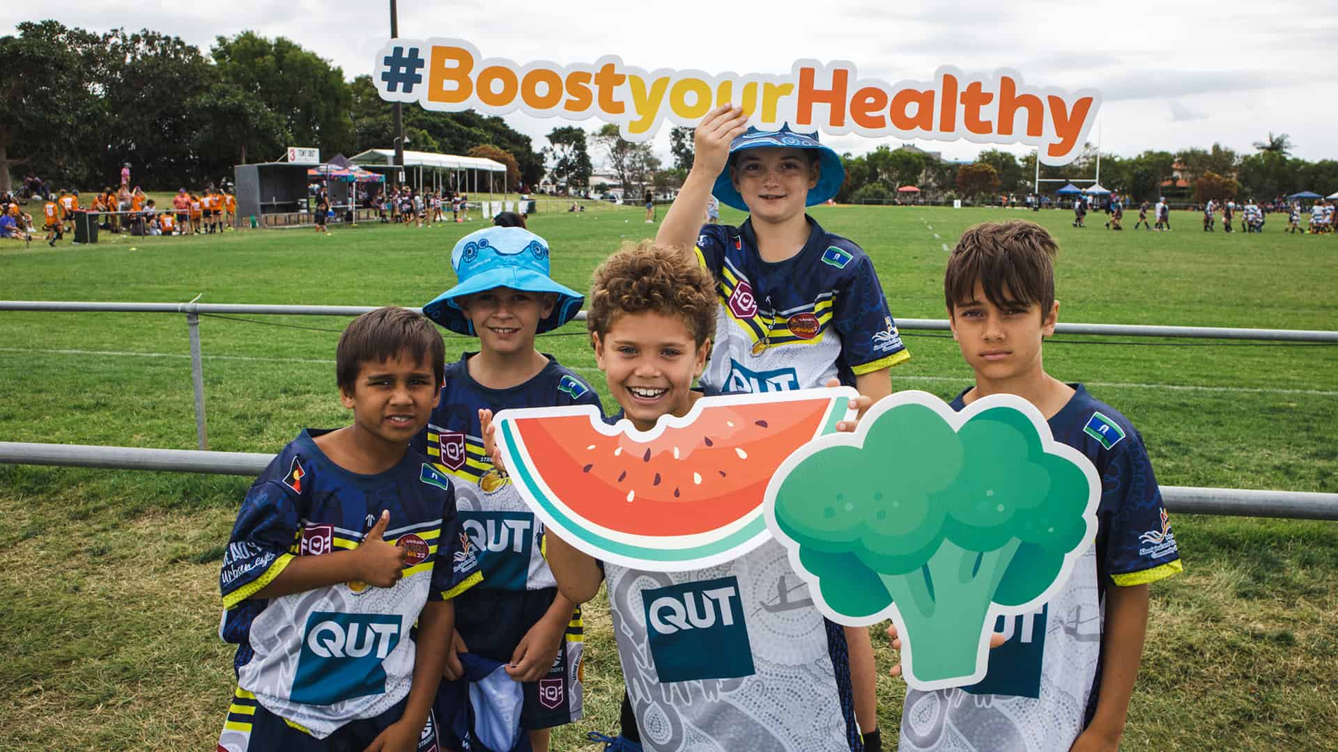 5 children on a sporting field are holding colourful signs in the shape of watermelon, broccoli and words that say #boostyourhealthy