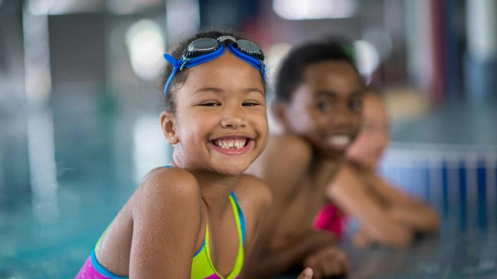 3 smiling children holding onto the edge of a swimming pool. One of the children is wearing swimming goggles on her head.