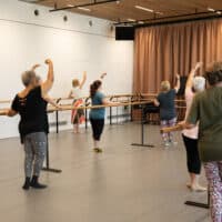 Seven middle-aged women practicing ballet holding onto a timber ballet pole in active wear.