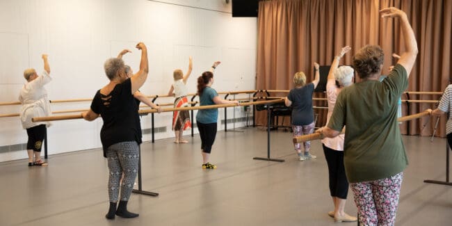 Seven middle-aged women practicing ballet holding onto a timber ballet pole in active wear.