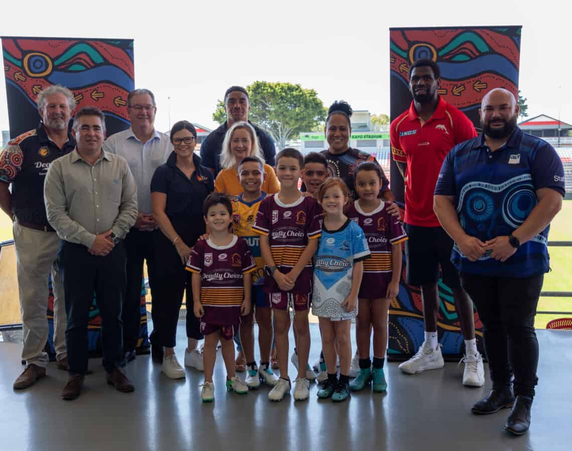 Group of leaders and six young boys wearing rugby league jerseys standing in the grandstand of a NRL stadium smiling side by side at the camera.