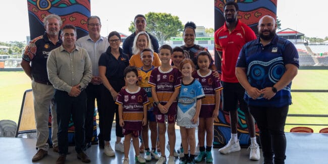 Group of leaders and six young boys wearing rugby league jerseys standing in the grandstand of a NRL stadium smiling side by side at the camera.