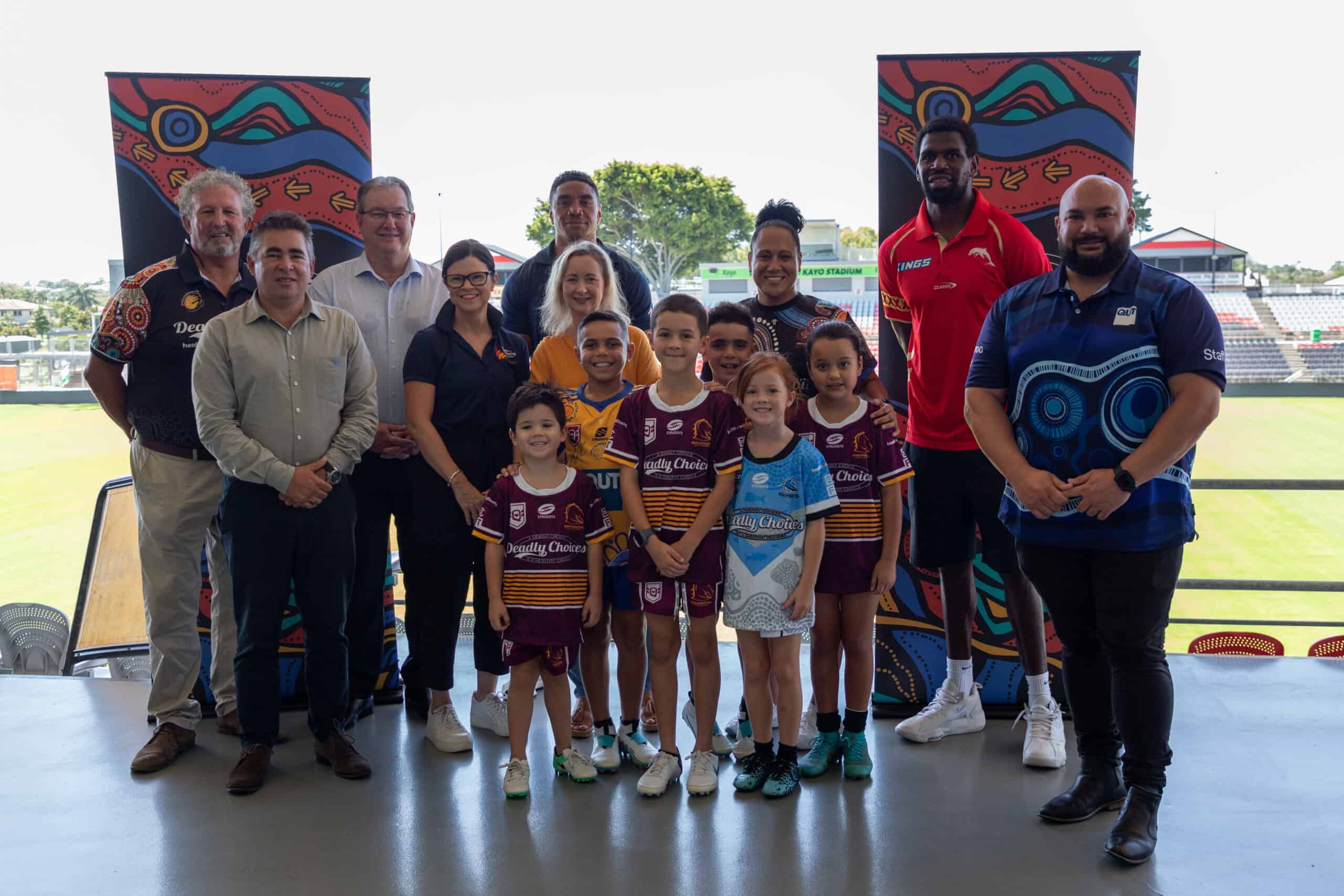 Group of leaders and six young boys wearing rugby league jerseys standing in the grandstand of a NRL stadium smiling side by side at the camera.