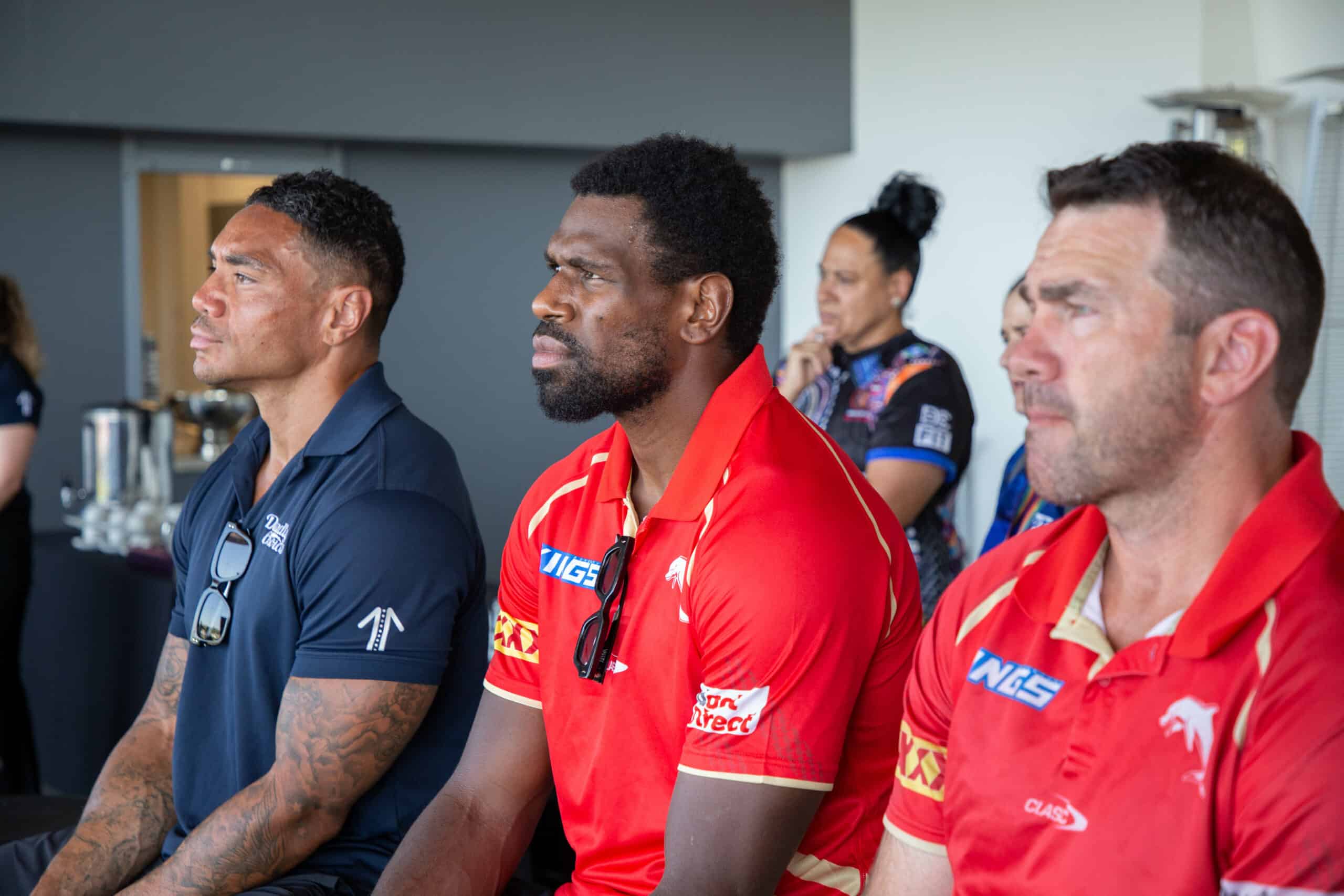 Three men side profile sitting in an audience listening to a speaker. Two men wear red, Redcliffe Dolphins jerseys and one wears a navy collared shirt.