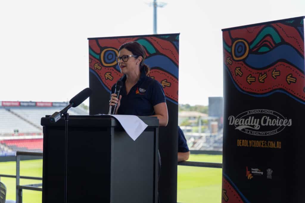 Robyn Littlewood (CEO Health and Wellbeing Queensland) standing behind a black plinth with a microphone. Two Deadly CHoices banners stand behind her. 