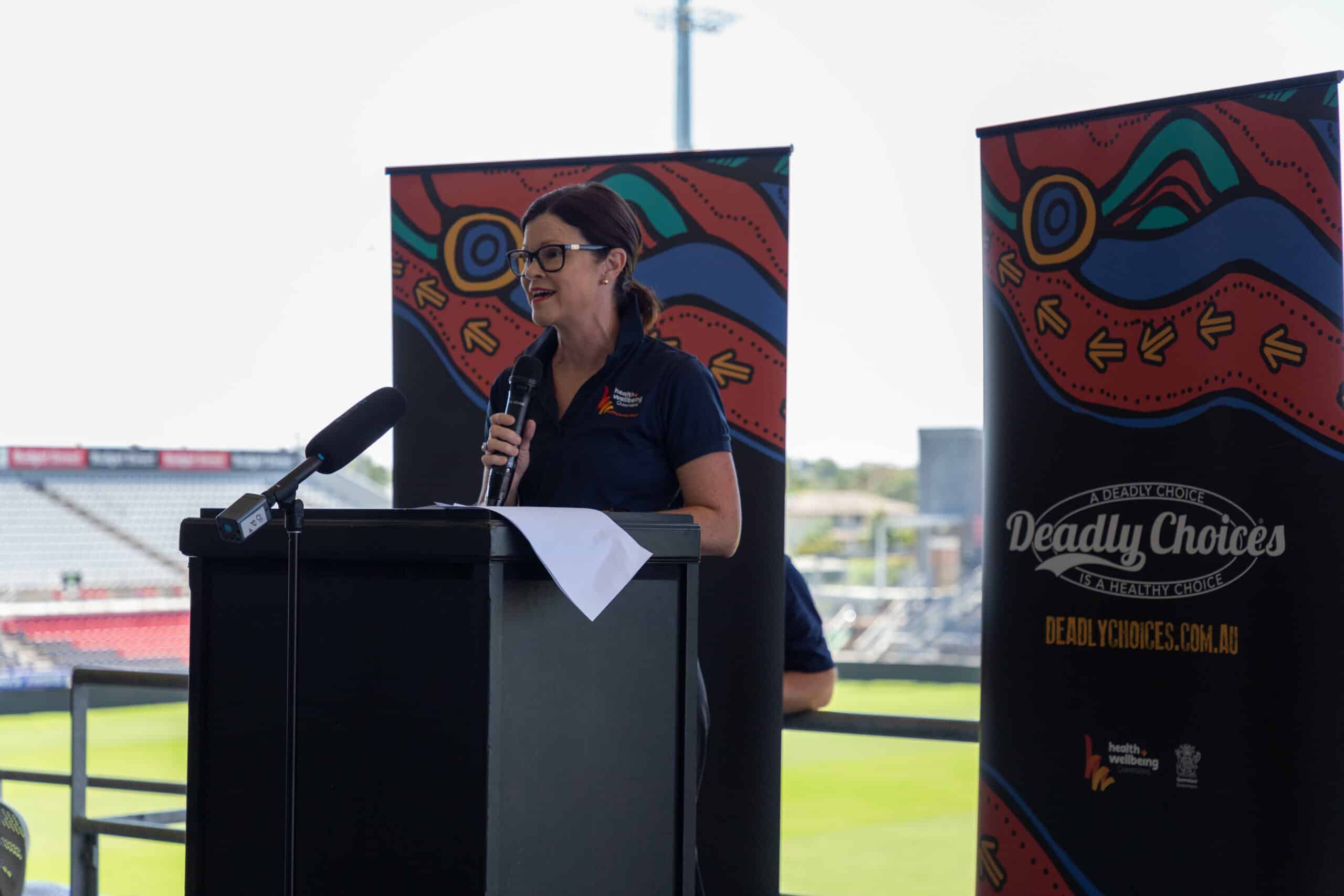 Robyn Littlewood (CEO Health and Wellbeing Queensland) standing behind a black plinth with a microphone. Two Deadly CHoices banners stand behind her. 