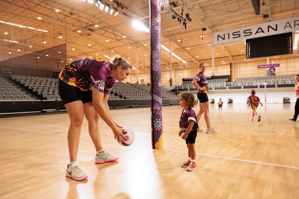 Donnell Wallam throwing a netball to a three year old young child on an indoor netball court. 