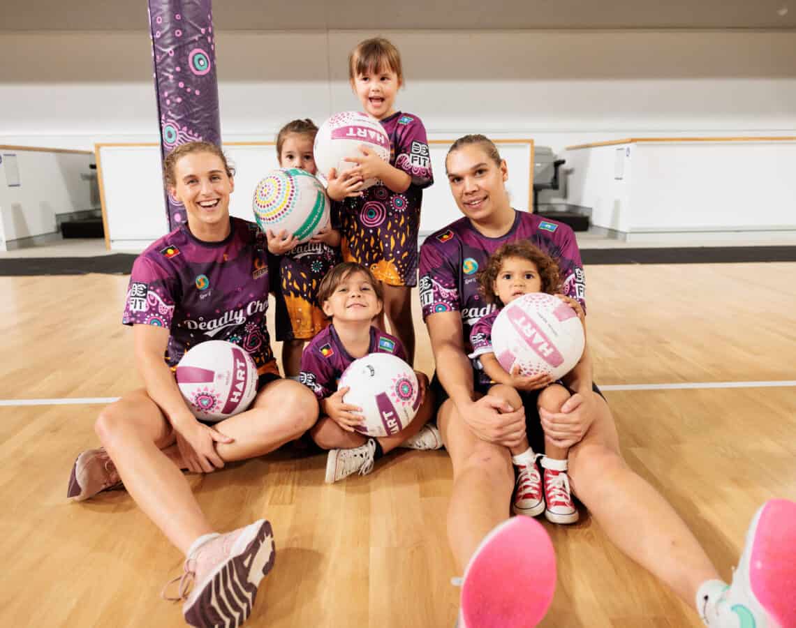 Two Firebirds players sitting on an indoor netball court with four young netball players under 5 years of age.