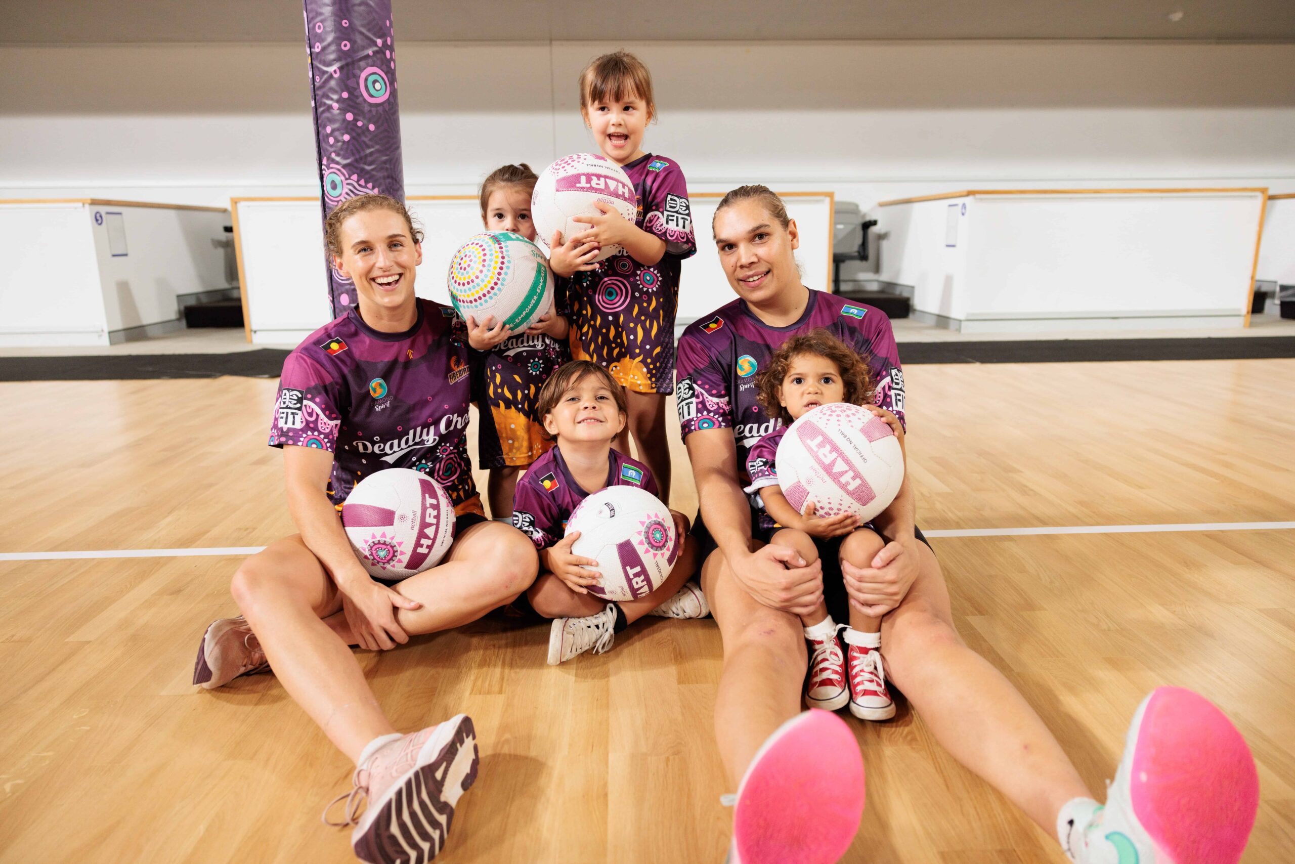 Two Firebirds players sitting on an indoor netball court with four young netball players under 5 years of age.