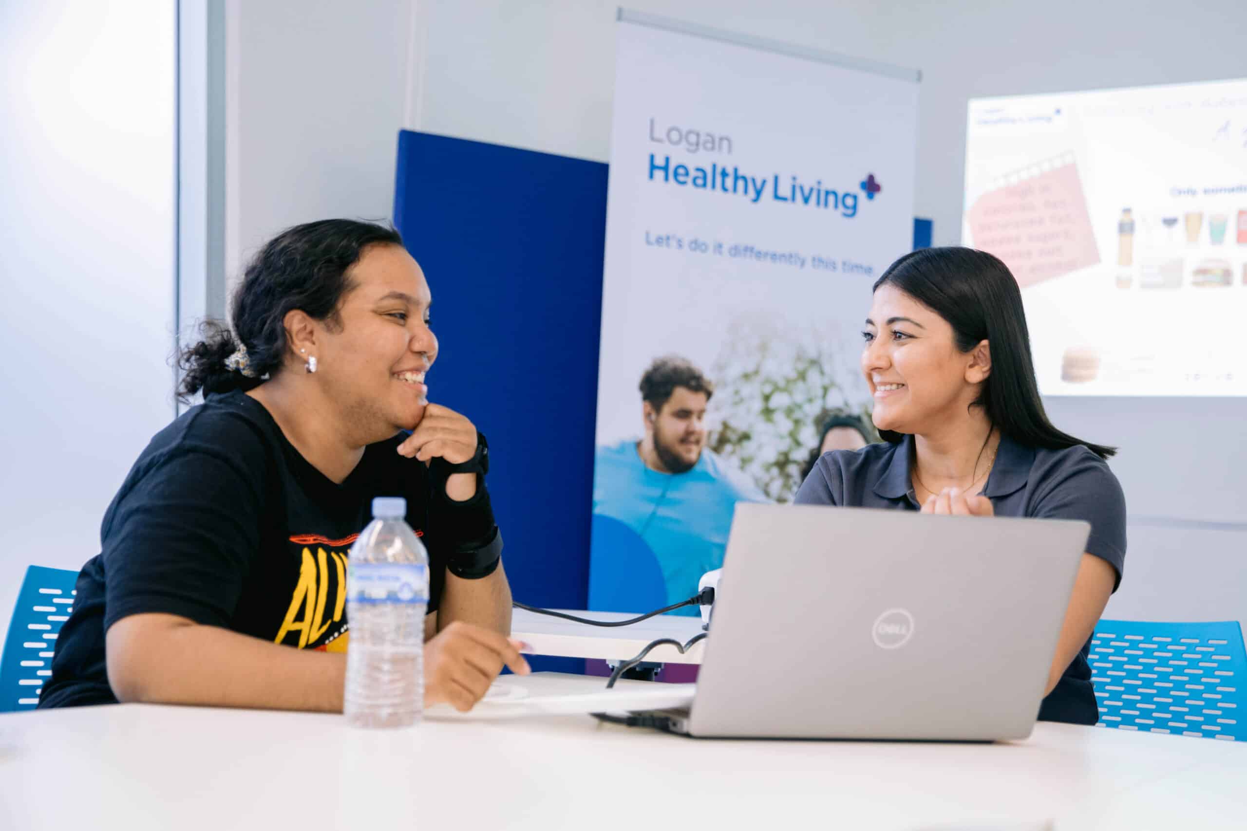 Two women sitting next to eachother at a laptop on a desk. Healthy Living pull up banner sits behind them.